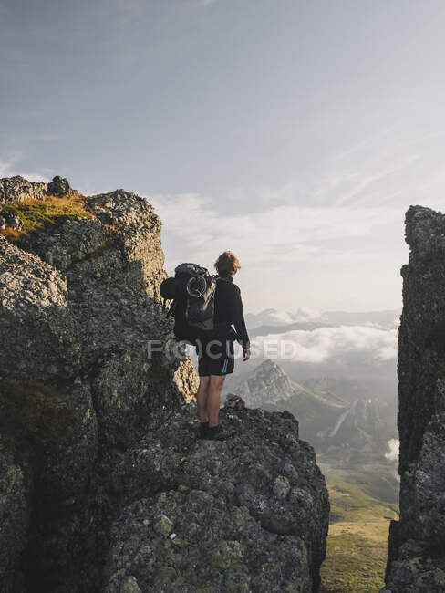Jeune homme avec sac à dos se tient à un sommet de falaise et regarde le paysage — Photo de stock