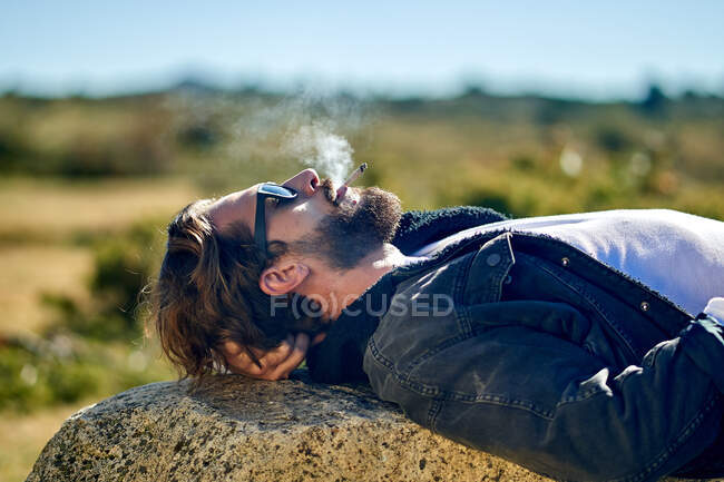 Porträt eines jungen tätowierten Mannes, der rauchend auf einem Felsen liegt — Stockfoto