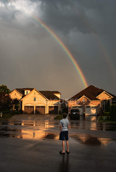 Boy standing in the street looking up at a rainbow in the sky. — Stock Photo
