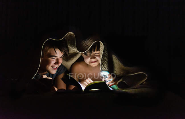 Brothers reading a book under a blanket by flashlight at night. — Stock Photo