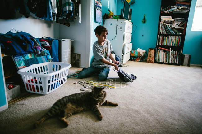 Niño en su dormitorio poniendo una camisa en una percha con su gato cerca - foto de stock