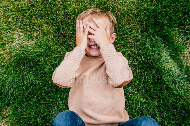 Happy young boy smiling and covering his face while laying in grass — Stock Photo