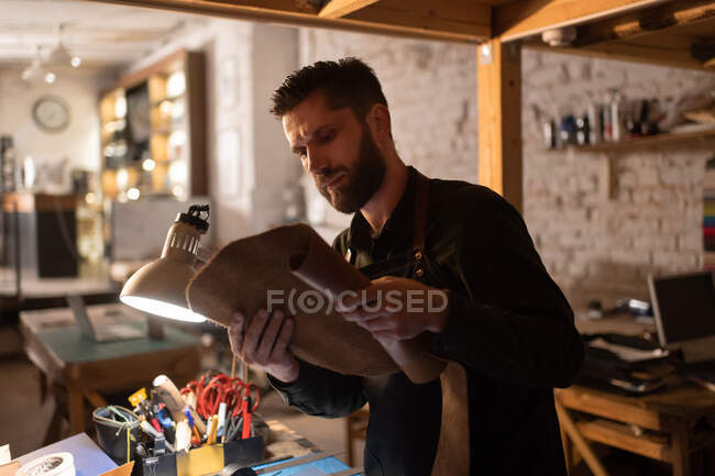 Bearded man in apron inspecting piece of leather while standing near illuminated table in home workshop and working — Stock Photo