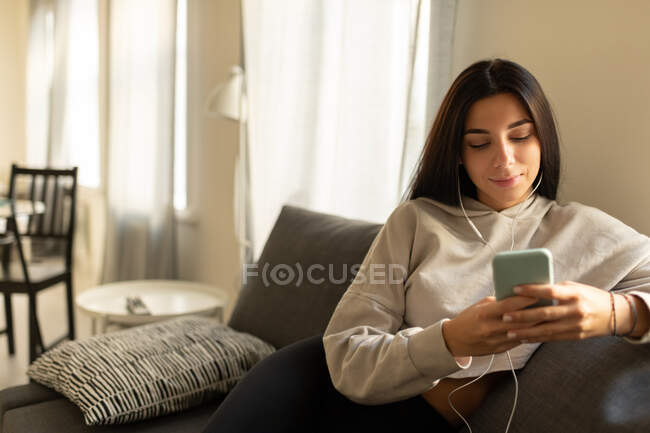 Задоволена молода брюнетка в навушниках і домашньому одязі переглядає смартфон, слухаючи музику, сидячи на дивані в кімнаті — стокове фото