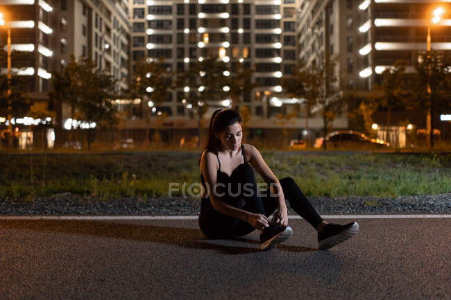 Mujer atlética joven en ropa deportiva sentada en la pista y atando cordones de zapatos mientras se entrena en el fondo urbano por la noche - foto de stock