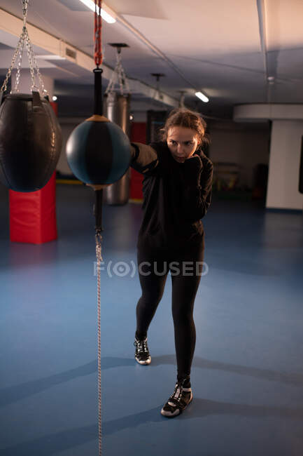 Повноцінне тіло підійде жінці в чорному спортивному одязі, яка б'є важкий мішок з джебом під час професійного тренування з боксу в сучасному спортзалі — стокове фото