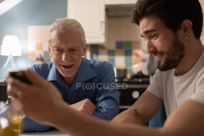 Fröhlicher junger Mann lächelt und zeigt erstaunten Vater lustige Fotos auf dem Smartphone, während er beim Familienessen in der gemütlichen Küche sitzt — Stockfoto
