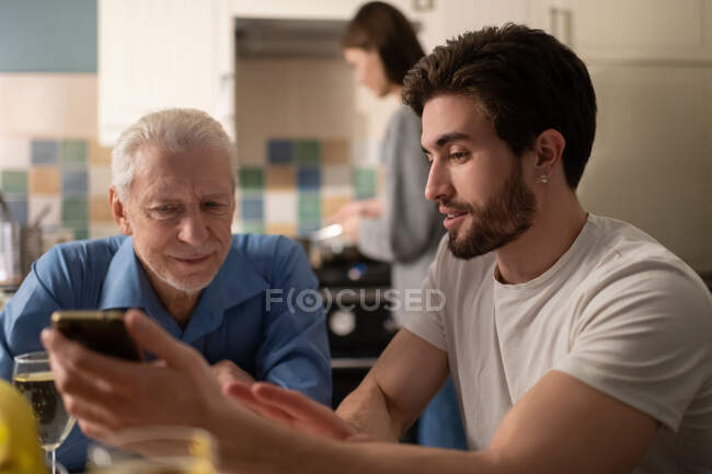 Joven hijo barbudo mostrando fotos en el teléfono inteligente a padre maduro interesado mientras se comunica cerca de la mujer cocinera durante la cena familiar en casa - foto de stock