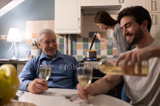 Cheerful mature man smiling and watching young male pouring wine while sitting near cooking woman before family dinner at home — Stock Photo