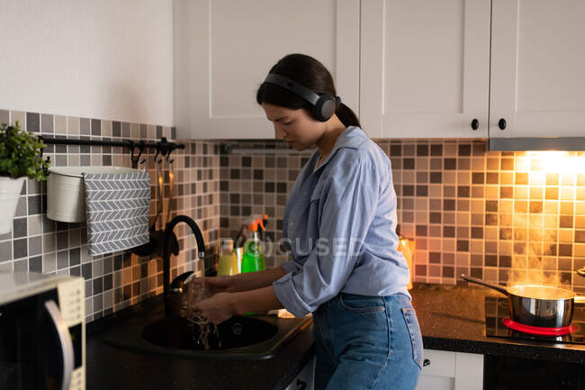 Vista lateral de la joven ama de casa en camisa azul y jeans escuchando música con auriculares inalámbricos y lavando platos en la cocina casera con agua hirviendo en la sartén en el fondo - foto de stock