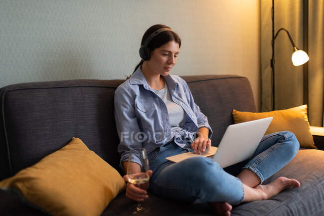 Femme décontractée détendue en chemise et en jeans se reposant à la maison après avoir travaillé dur assis sur un canapé avec des oreillers boire du vin et regarder un film sur ordinateur portable avec écouteurs sans fil — Photo de stock