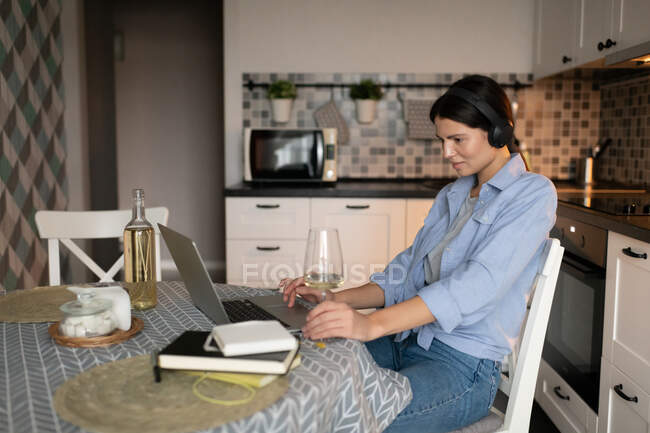 Side view of modern woman in casual clothes enjoying free time and browsing internet on laptop while sitting at table in kitchen drinking wine and listening to music with wireless headphones — Stock Photo