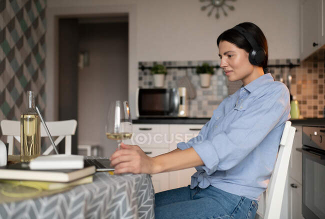 Vista lateral de la joven mujer casual en auriculares con computadora portátil y beber vino mientras está sentada en la mesa en la cocina y descansando después de las tareas domésticas - foto de stock