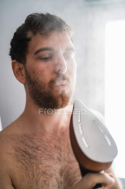 Hombre soplando en el hierro caliente emitiendo vapor mientras hace las tareas domésticas - foto de stock