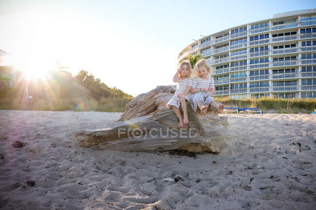 Zwei Mädchen, die auf Treibholz am Strand sitzen und die Sonne hinter sich haben — Stockfoto