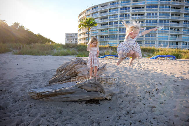 Deux filles sur le bois flotté sautant dans le sable sur la plage — Photo de stock