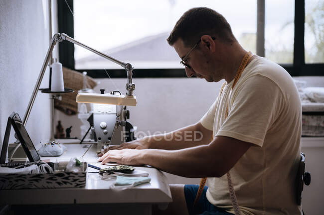 Junger Mann schaut einer Anleitung zu, während er mit der Nähmaschine Masken herstellt — Stockfoto