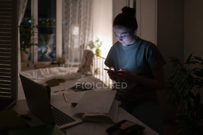 Estudiante verificando correo electrónico en teléfono móvil en habitación oscura durante el aprendizaje a distancia - foto de stock
