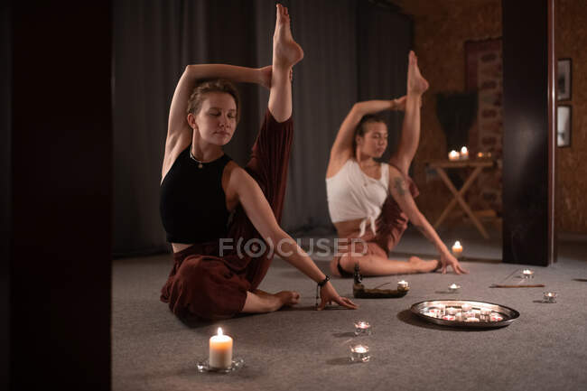 Slim amiche di sesso femminile che si estende in posa Krauncasana mentre seduto vicino alle candele — Foto stock