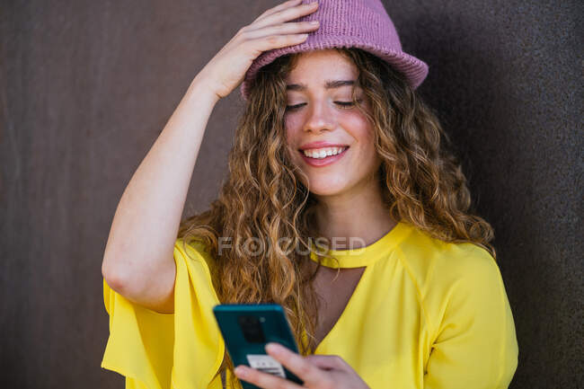 Задоволена жінка регулює стильний капелюх під час використання смартфона — стокове фото