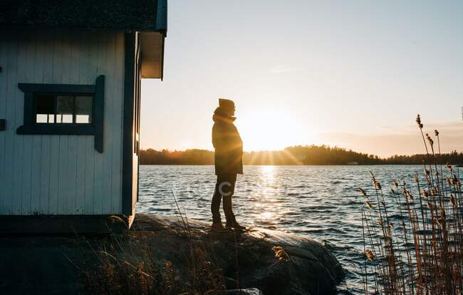 Людина стояла на скелі біля дерев'яної хатини в Швеції на заході сонця. — стокове фото