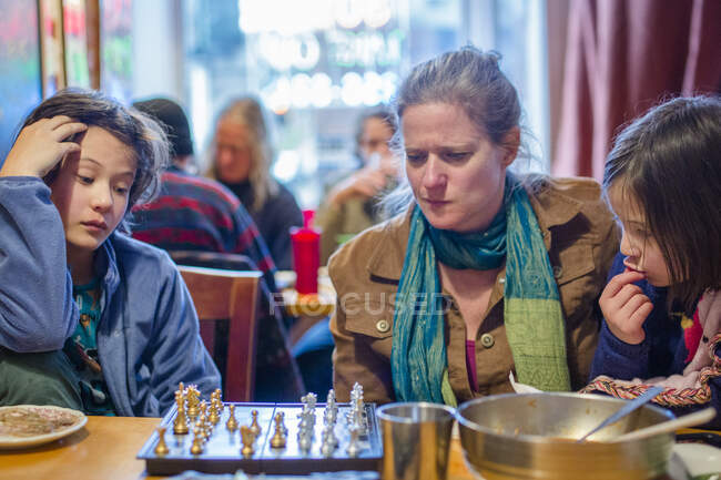 A woman and children sit together in restaurant studying chess board — Stock Photo