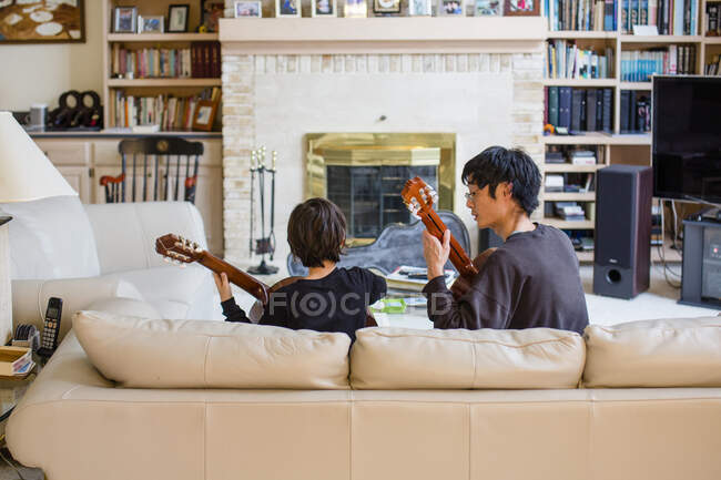 A father and son sit together on couch playing classical guitar music — Stock Photo