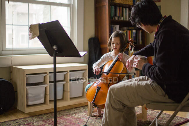 A father helps little girl practice cello at home by window — Stock Photo