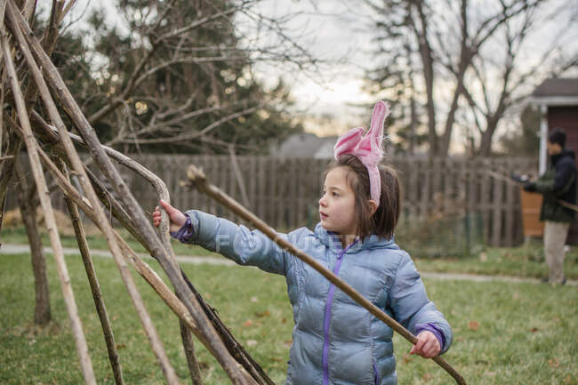 Uma menina em orelhas de coelho e casaco constrói forte com o pai no quintal — Fotografia de Stock
