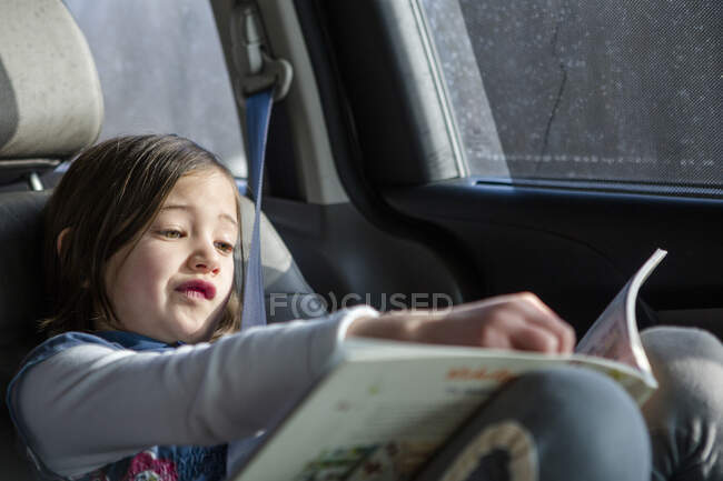 A cute little girl sits in a carseat in sunlight studying a book — Stock Photo