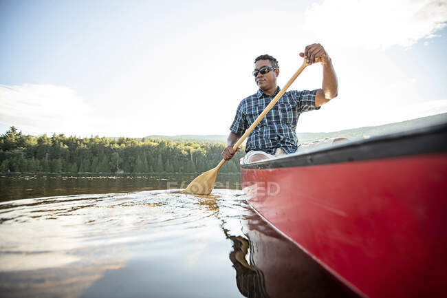 Sonriente hombre afroamericano en canoa roja sumerge remo en el lago en Maine - foto de stock
