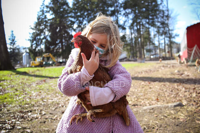 Chica sosteniendo pollo en una granja usando una máscara - foto de stock