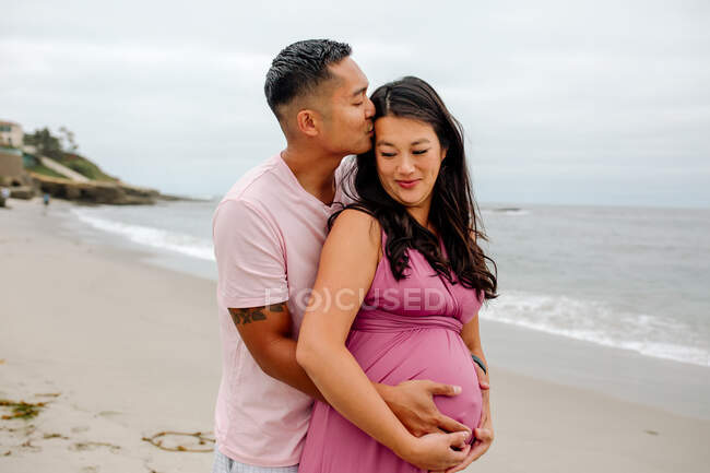Любящий азиатский муж целует голову улыбающейся беременной жены на пляже — стоковое фото