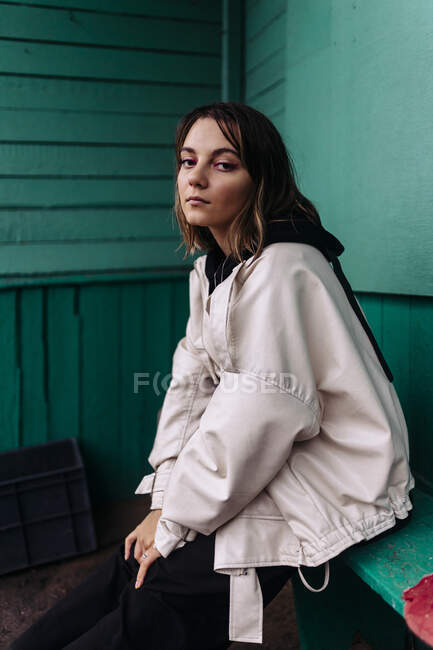 Donna felice con una giacca bianca seduta su una panchina vicino a un muro verde — Foto stock