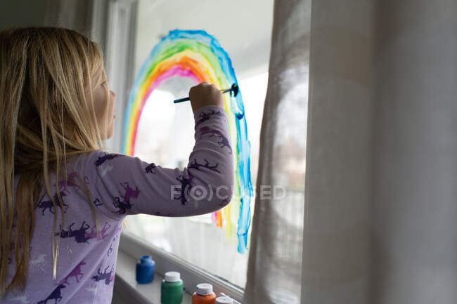 Blond girl painting rainbow on interior window in house — Stock Photo