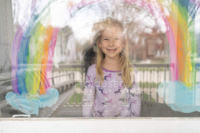 Sourire blond jeune fille sous arc-en-ciel peint sur la fenêtre — Photo de stock