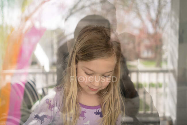 Outside in view of blond girl painting rainbow on window — Stock Photo