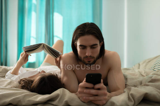 Donna che legge il libro sul letto vicino all'uomo usando lo smartphone mentre riposa al mattino a casa — Foto stock