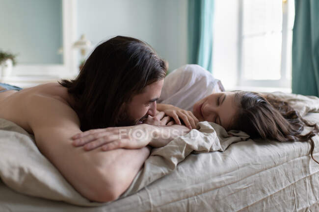 Jovem e mulher sorrindo e olhando um para o outro enquanto descansam na cama em casa — Fotografia de Stock