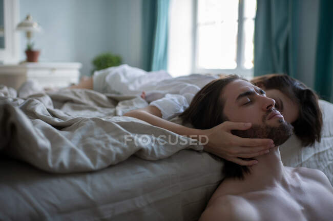 Jeune femme reposant sur le lit et caressant homme les yeux fermés dans la chambre sombre — Photo de stock