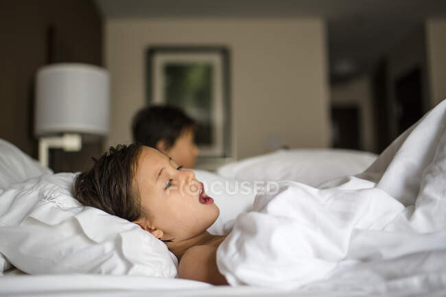 A little girl lays in a hotel room bed singing brother in background — Stock Photo
