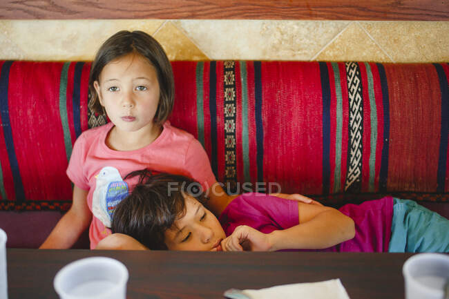Una niña pequeña se sienta en la mesa de un restaurante la cabeza de su hermano en su regazo - foto de stock