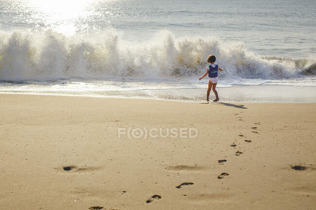 Uma menina em colete salva-vidas fica na frente da onda que se aproxima na praia — Fotografia de Stock