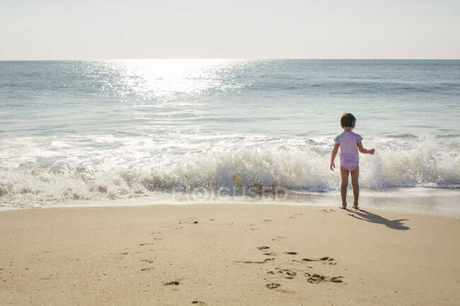Une petite fille se tient au bord du rivage avec une vague venant en sens inverse — Photo de stock