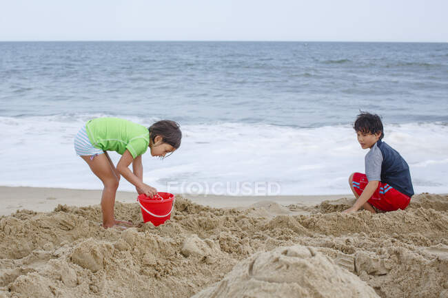 Two children play together with a red bucket on sandy beach by ocean — Stock Photo