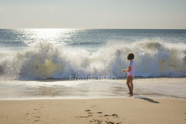 A small girl stands at edge of ocean watching wave break onto beach — Stock Photo