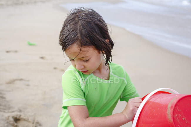 Маленькая девочка с мокрыми песчаными волосами играет с ведром на песчаном пляже — стоковое фото
