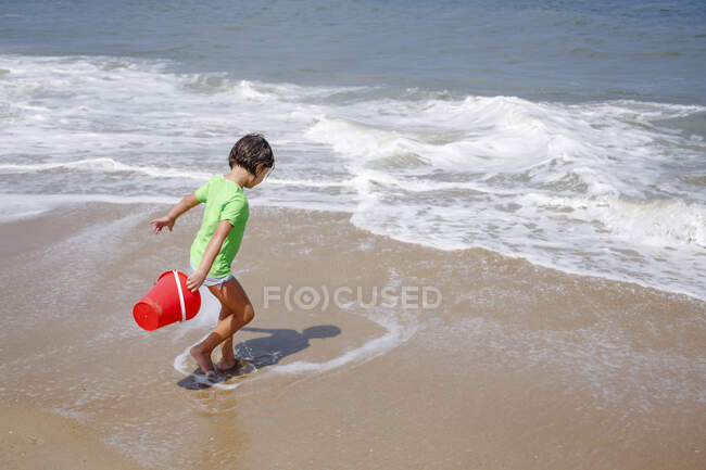 Маленькая девочка с ведром играет в приливе на краю песчаного пляжа — стоковое фото