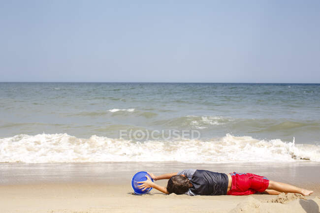 A boy lays on a beach with a blue bucket waiting for oncoming wave — Stock Photo