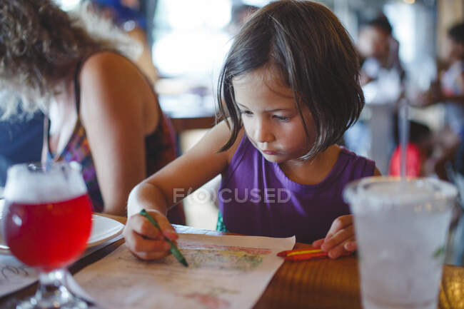 Una niña se sienta en una mesa de restaurante para colorear con lápices de colores - foto de stock